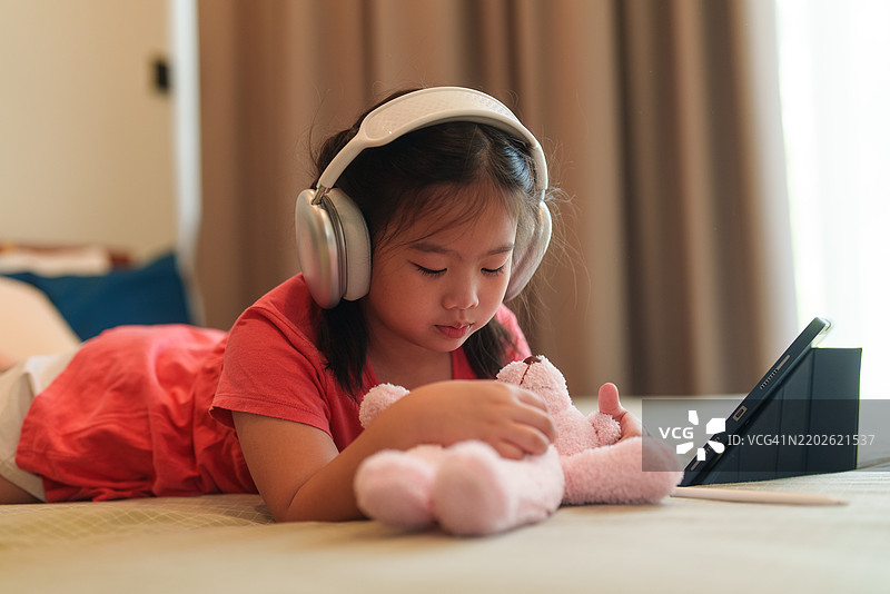 一个可爱的女孩在周末的卧室里，戴着耳机，使用平板电脑在床上画画和做作业，玩得很开心。图片素材