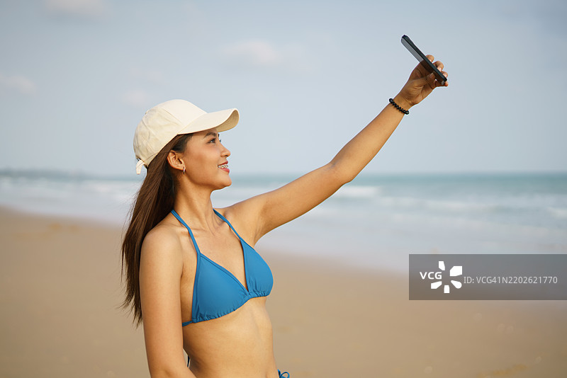 美丽的年轻亚洲女性在海滩上穿着比基尼自拍，手持智能手机。夏季海滩概念。图片素材