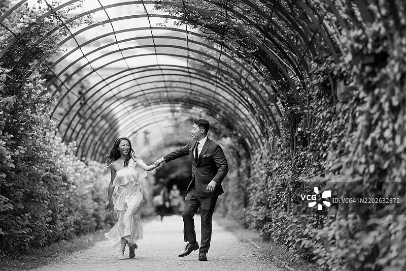 一对情侣在阳光明媚的花园中盛开的拱门下摆姿势。阳光营造出一种神奇的氛围，非常适合浪漫。图片素材