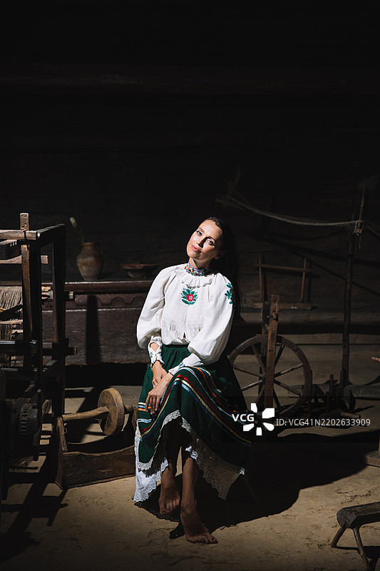 一位身着传统服饰的女性坐在一个充满编织工具和陶器的乡村工作室里，柔和的光线洒在周围。图片素材