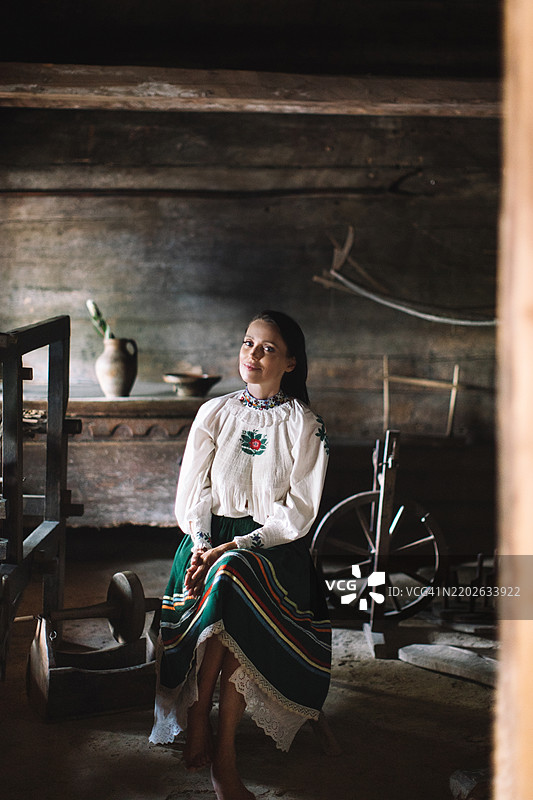 一位身着传统服饰的女性坐在一个充满编织工具和陶器的乡村作坊里，柔和的光线洒在周围。图片素材