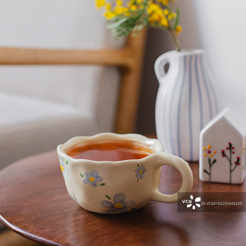 一杯热茶，一个小陶瓷房子，花瓶里的含羞草，放在温馨的椅子上的木咖啡桌上。舒适的氛围。你好，春天。复活节或母亲节的概念。图片素材