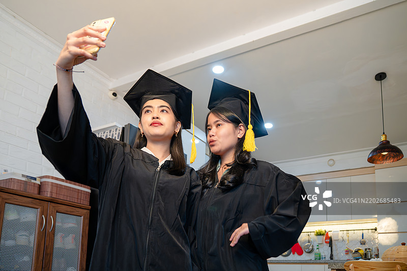 两位年轻女性穿着毕业袍和学位帽，通过视频通话向亲戚或朋友问好。图片素材