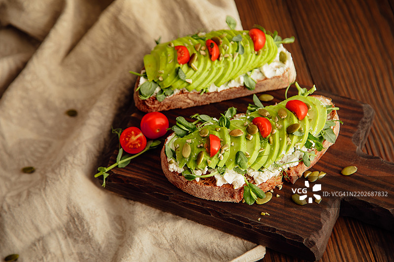 两片乡村面包吐司，上面放有豌豆芽、奶油奶酪、鳄梨、南瓜子和番茄。图片素材
