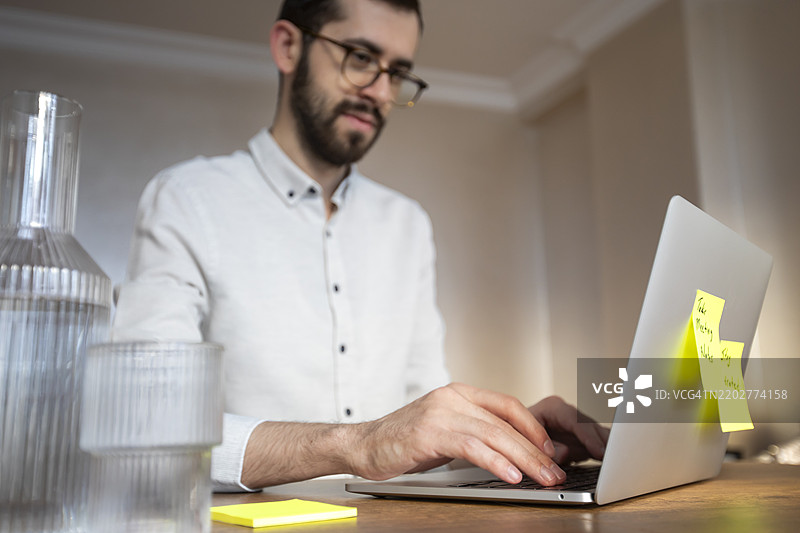 一位穿着白衬衫的男士正在使用笔记本电脑，桌上贴着两个便签。一个写着“记录会议笔记”，另一个写着“保持水分 :)”。桌上还有一个水瓶和一盆植物。图片素材