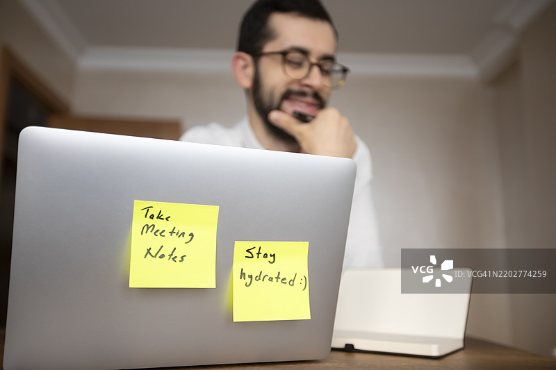 一位穿着白衬衫的男士正在使用笔记本电脑，桌上贴着两个便签。一个写着“记录会议笔记”，另一个写着“保持水分 :)”。桌上还有一瓶水和一盆植物。图片素材
