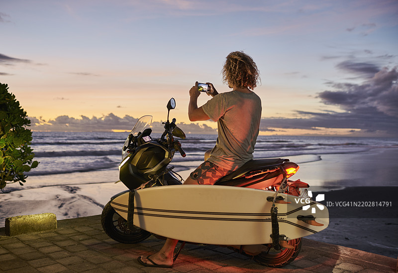 男性冲浪者坐在摩托车上，面朝巴厘岛海滩，使用智能手机拍摄美丽的日落。图片素材