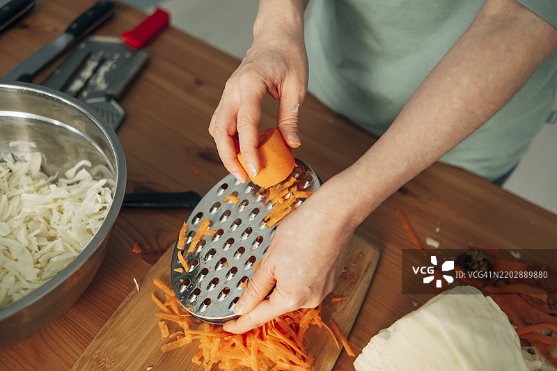 女人使用特殊的擦菜器擦胡萝卜。腌制白菜的准备过程。发酵产品。女人的手特写，俯视图。图片素材