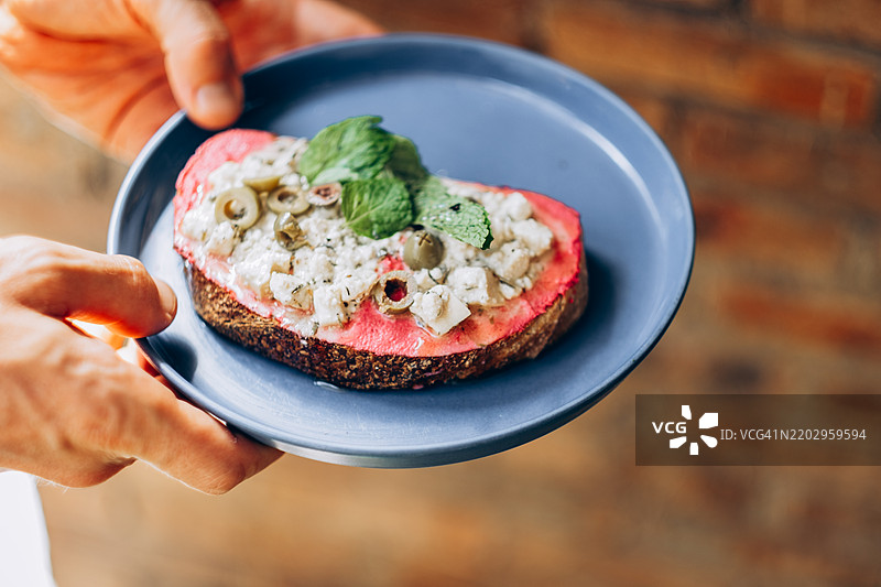 男人的手拿着一个蓝色盘子，上面放着一片涂有粉色甜菜根鹰嘴豆泥的吐司，顶部装饰有素食羊奶酪、绿橄榄和香草，背景是砖墙。图片素材