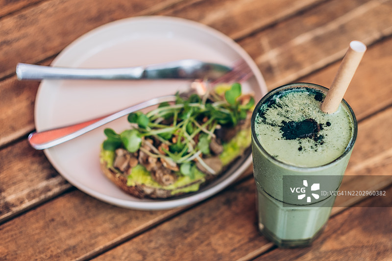 盘子里有鳄梨吐司，上面放着炒香菇和微型绿叶菜，还有一杯绿色碱性果昔，配有可生物降解的吸管，摆放在木桌上。图片素材