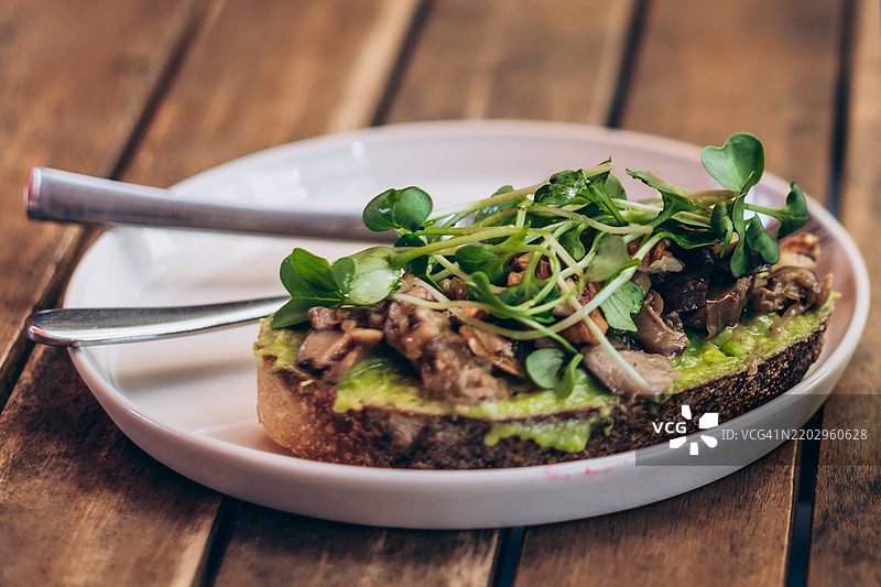 盘子里放着鳄梨吐司，上面点缀着炒香菇和微型绿叶菜。两把银色刀具静静地放在盘子上。木质桌子营造出温馨的用餐氛围。图片素材