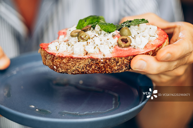 男人手持一片涂有粉色豆腐酱的吐司，上面放有素食羊奶酪、绿橄榄和新鲜薄荷。吐司放在一个蓝色盘子上，旁边有少许橄榄油。图片素材