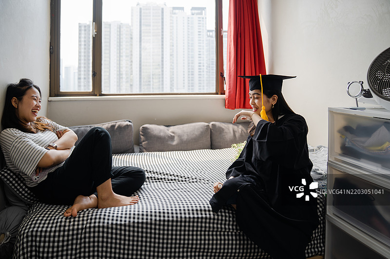 微笑的女性穿着毕业袍，在宿舍里，洋溢着快乐，帮助系上庆祝的丝带。图片素材