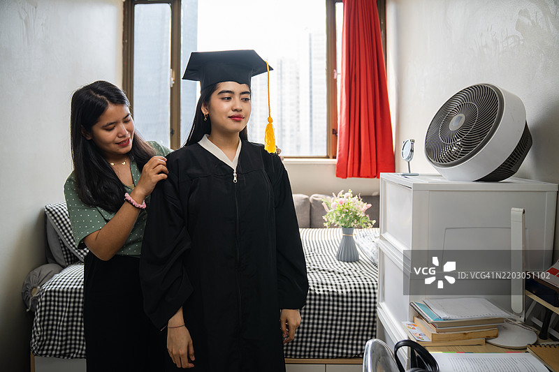 微笑的女性穿着毕业袍，在宿舍里，洋溢着快乐，帮助系上庆祝的丝带。图片素材