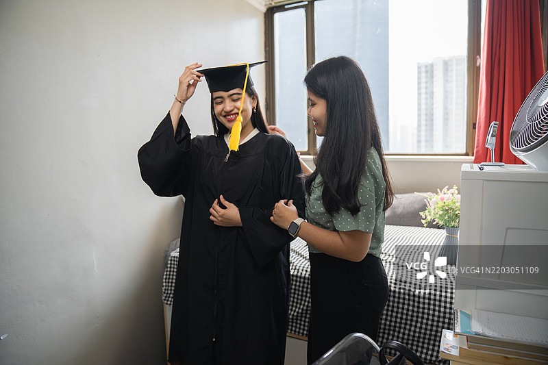 微笑的女性穿着毕业袍，在宿舍里，洋溢着快乐，帮助系上庆祝用的丝带。图片素材