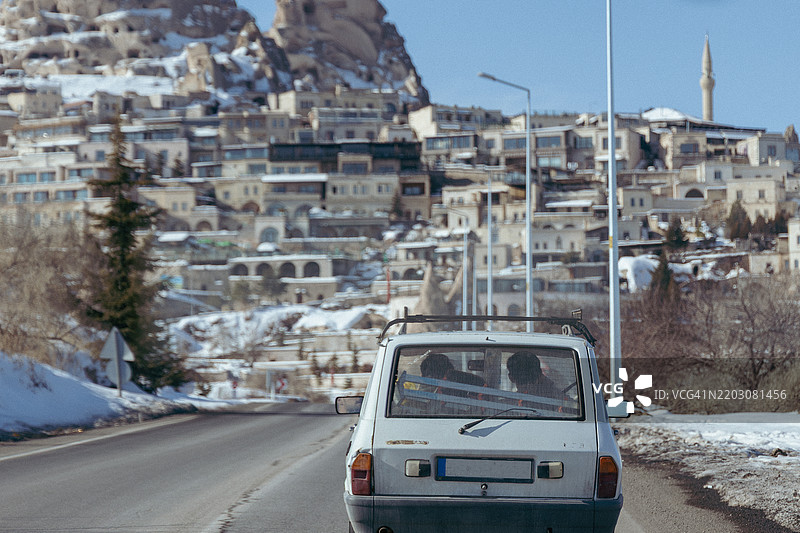 复古汽车沿着通往鸽子谷的道路行驶，背景是被雪覆盖的乌奇希萨尔城堡，土耳其中部安纳托利亚地区的美丽风景。图片素材