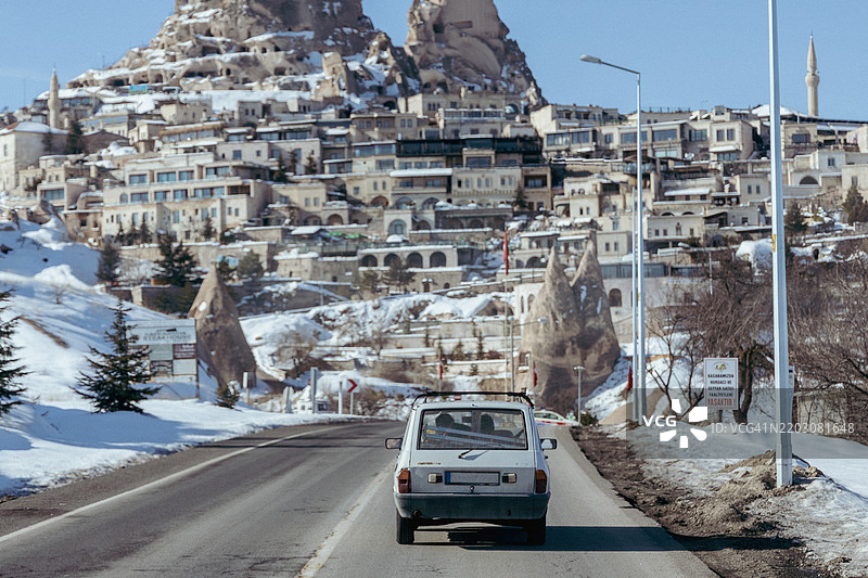 复古汽车沿着通往鸽子谷的道路行驶，背景是被雪覆盖的乌奇希萨尔城堡，中央安纳托利亚，土耳其。图片素材
