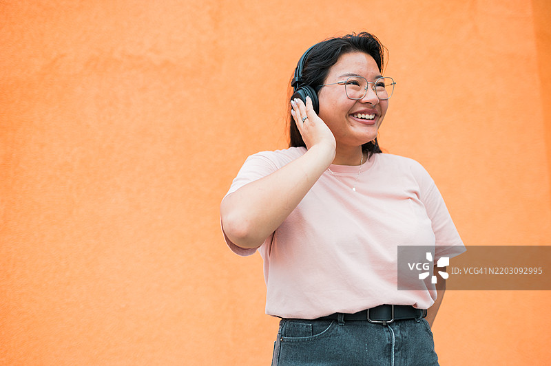 一位有趣的微笑韩国年轻女性在橙色墙壁前戴着耳机享受音乐，旁边留有空白空间。图片素材