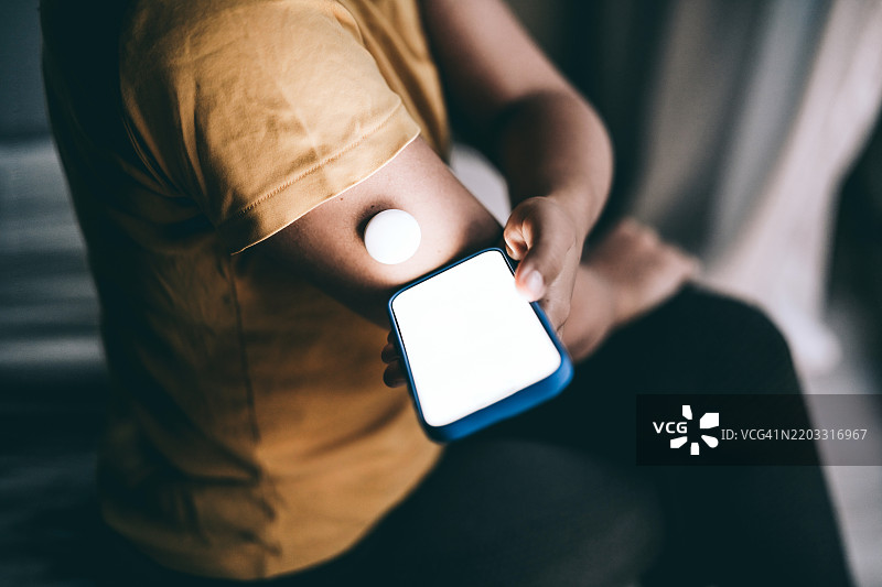 一个无法辨认的人正在使用血糖监测仪，查看手机应用以获取更多血糖水平数据。图片素材