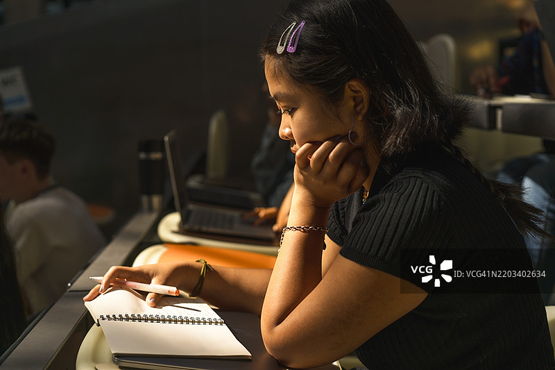 年轻女学生坐在大学讲堂里，手托着下巴阅读书籍。图片素材
