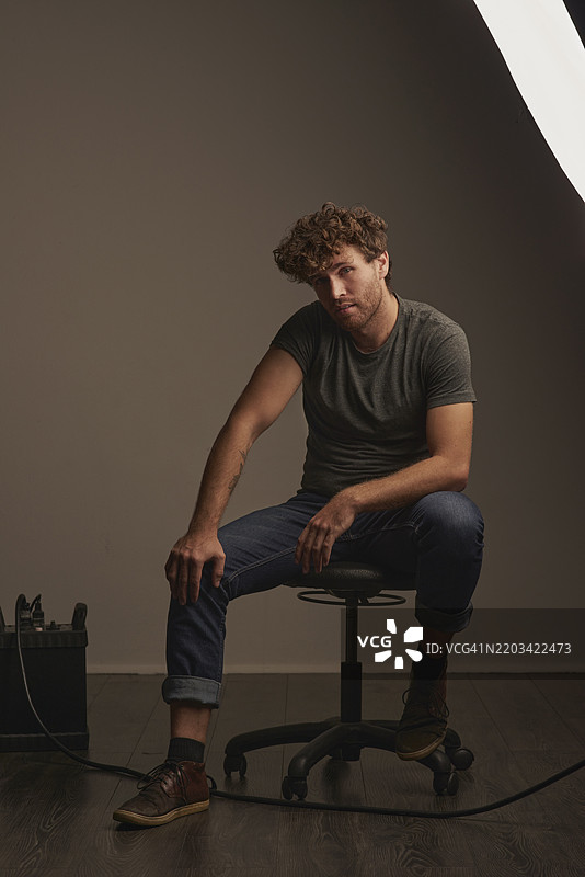 肖像，摄影棚中一位英俊男士坐在椅子上，背景为灰色，展现时尚。摄影、幕后和设备，年轻人在拍摄期间坐在幕后。图片素材