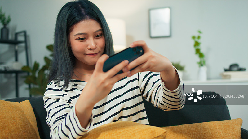 年轻的亚洲女性在家沙发上放松，玩手机在线游戏，享受休闲活动。图片素材