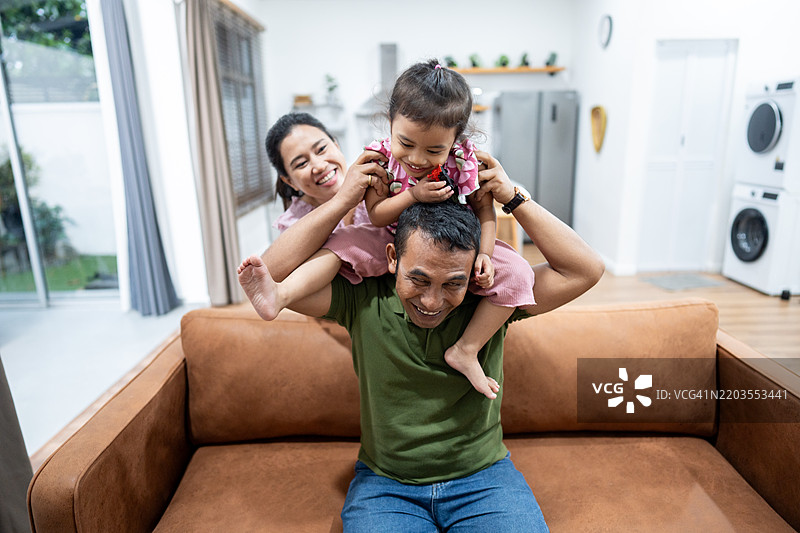 一个快乐的家庭时刻，父亲给女儿背着，母亲在家中温暖地微笑。图片素材