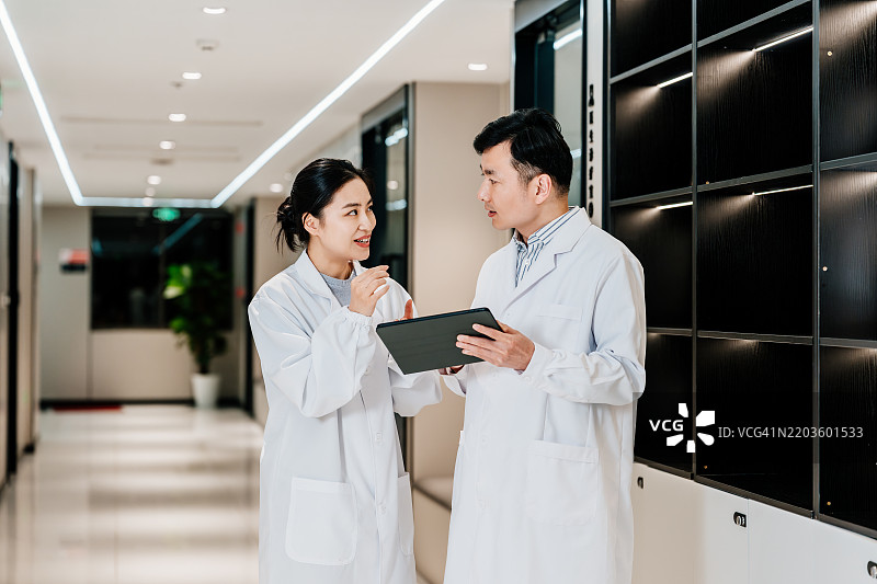 两位亚洲医生在医院走廊里站着讨论数字平板上的工作。图片素材