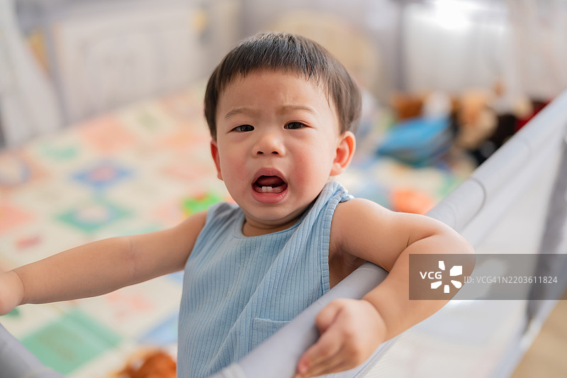 亚洲婴儿男孩站在婴儿床里，面露悲伤的表情，哭泣。这个宝宝在家里白天哭泣并呼唤妈妈，展现了家庭生活中的婴儿情景。图片素材