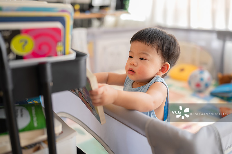 可爱的小男孩学习站立，手抓着围栏，专注地试图从书架上拿一本书。图片素材