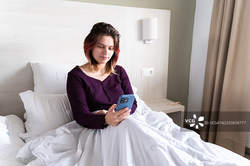 年轻女性在床上使用智能手机：早晨第一件事就是查看社交媒体图片素材