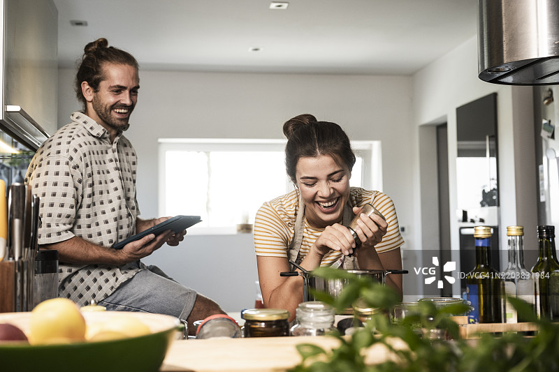 快乐的女人在家厨房里做饭，旁边的男人坐着用平板电脑。图片素材