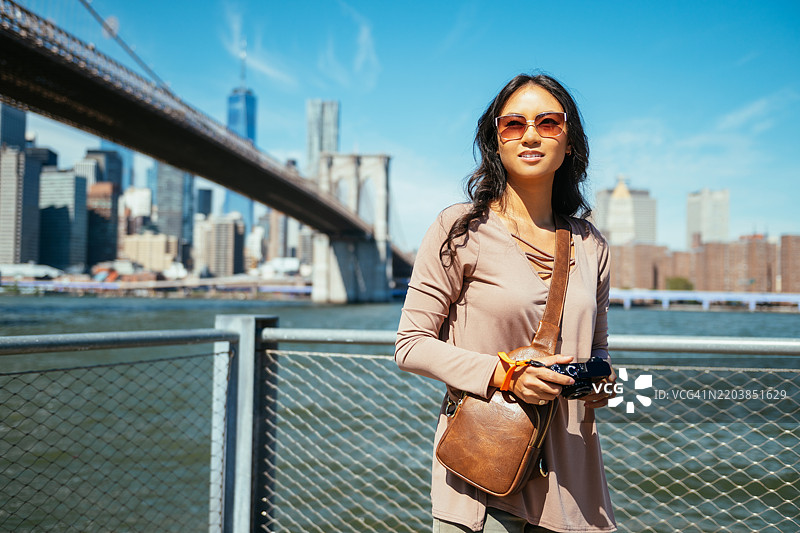 美丽的亚洲女性游客在纽约市，手持小型相机，站在曼哈顿天际线前。图片素材