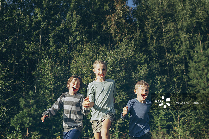 快乐的女孩在阳光明媚的日子里与兄弟们在树木前奔跑图片素材