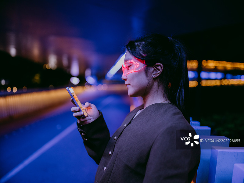 年轻的亚洲女商人戴着发光眼镜，站在发光的人行天桥下，正在使用她的智能手机。图片素材