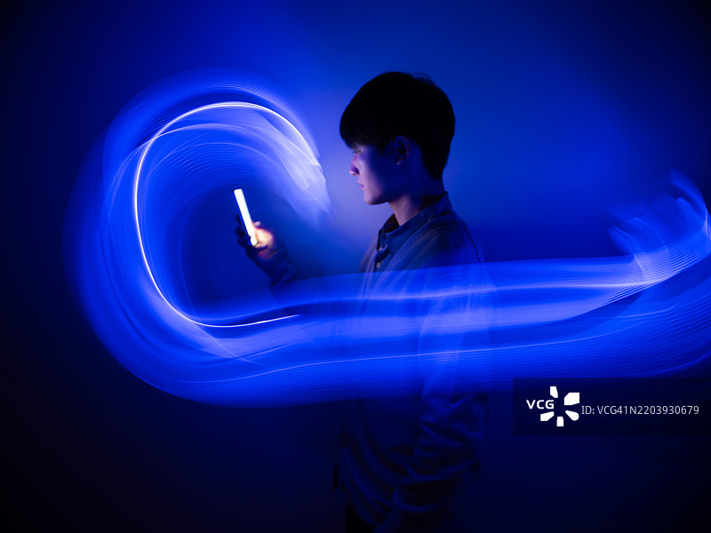 一名年轻男子在流动的光轨中使用智能手机。仿佛置身于梦与现实之间的世界。人工智能概念，LED灯的长时间曝光。图片素材