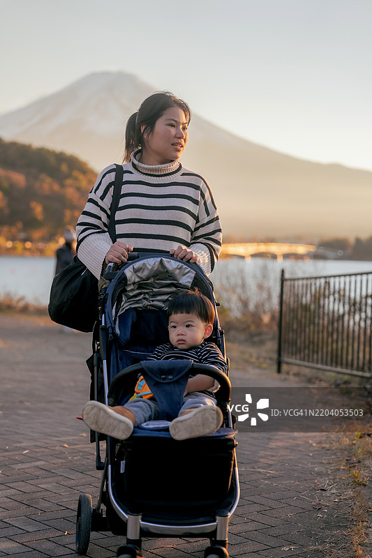 母亲推着婴儿车与家人在湖边散步，享受日落时刻，沿着河湖边走。周末旅行。图片素材