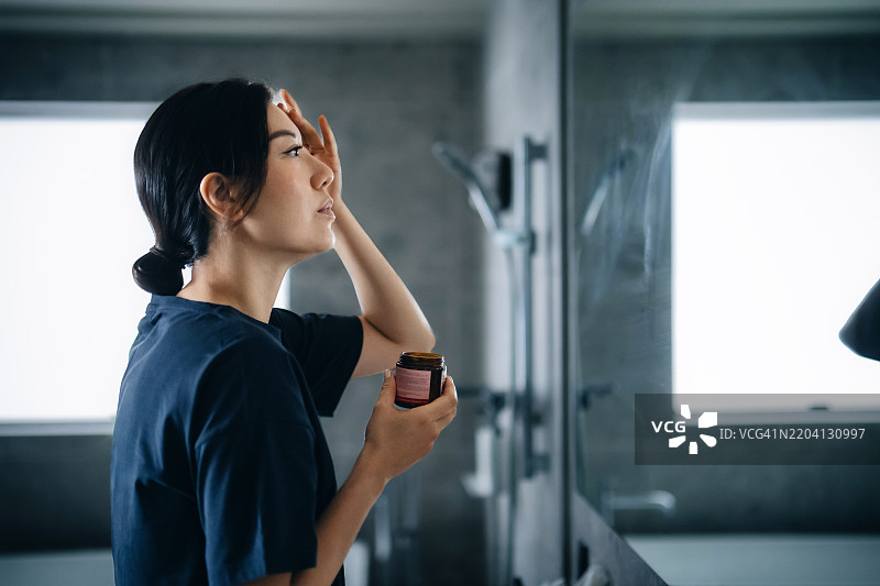 年轻亚洲女性侧面站在浴室镜子前，正在脸上涂抹面霜。家庭护理、美容、护肤和健康生活方式的概念。图片素材