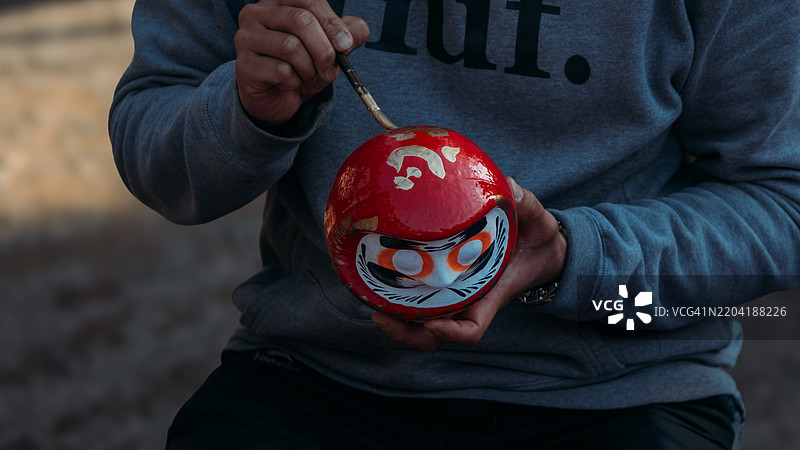 深大寺达摩娃娃节是东京调布市深大寺举办的一个受欢迎的春季节日，拥有超过300年的历史。画面展示了游客在达摩娃娃上写下愿望，祈求好运。图片素材