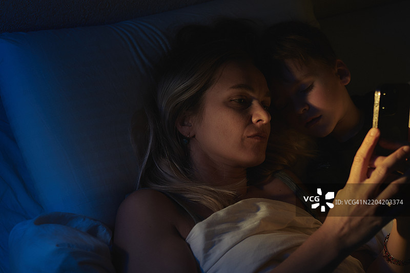 母亲和儿子在床上夜晚放松，一起翻看智能手机图片素材