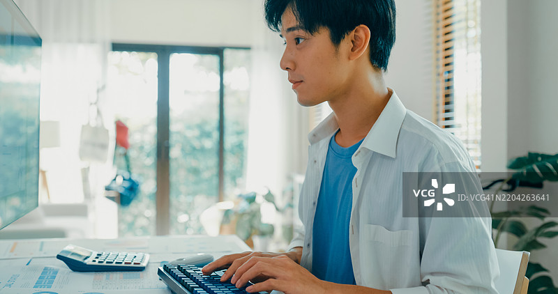 年轻亚洲男性在家工作，近距离观察他在电脑屏幕上分析财务数据，反映出现代家庭办公室的布局和远程工作的状态。远程工作与财务。图片素材