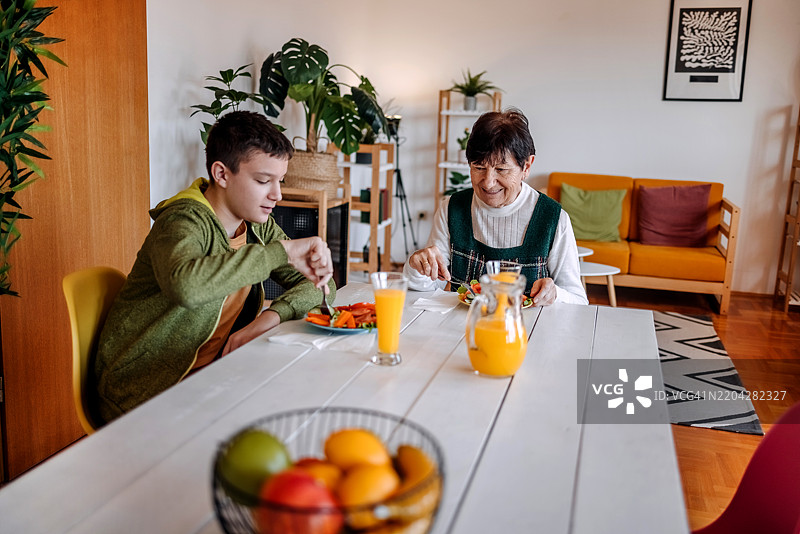 少年男孩在家中与祖母共度时光，享用健康早餐，坐在餐桌上写字。图片素材
