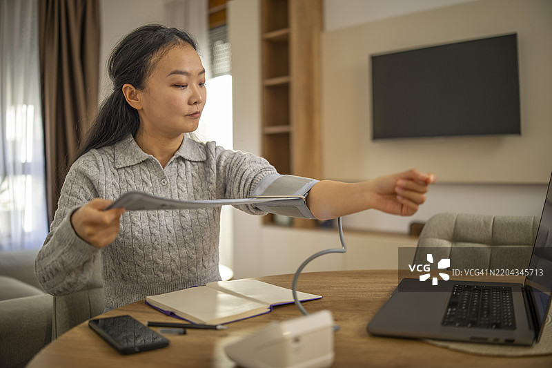 可爱的亚洲女性在家中的客厅桌子旁戴上数字血压计，然后检查血压。图片素材