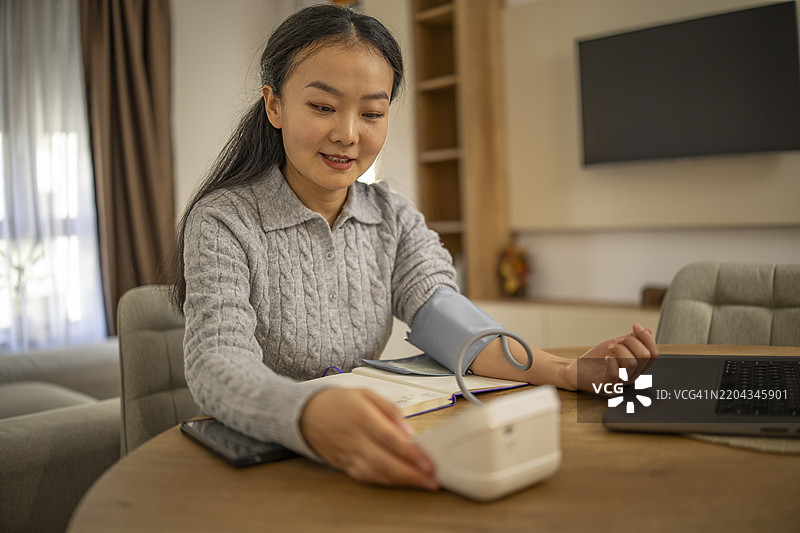 可爱的亚洲女性坐在家中的客厅桌子旁，用数字血压计测量血压和心率。图片素材