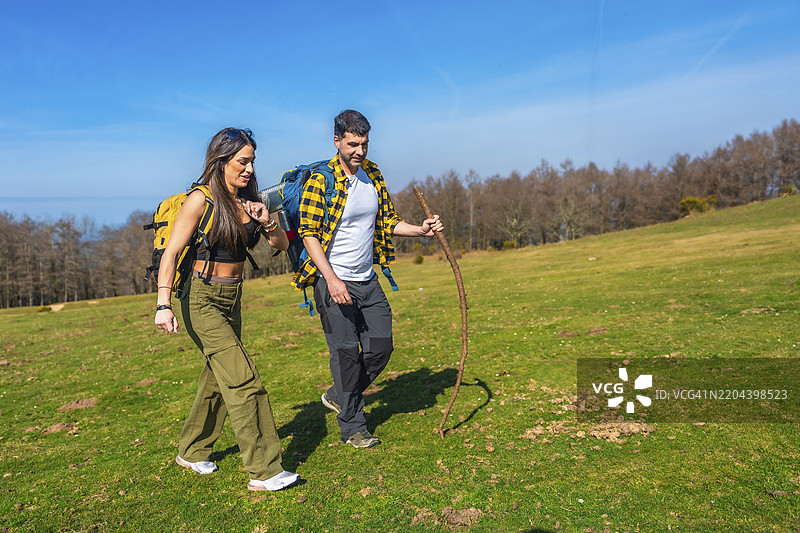 年轻情侣手牵手走过生机勃勃的绿草地，背着背包，手持登山杖，在阳光明媚的天空下。图片素材