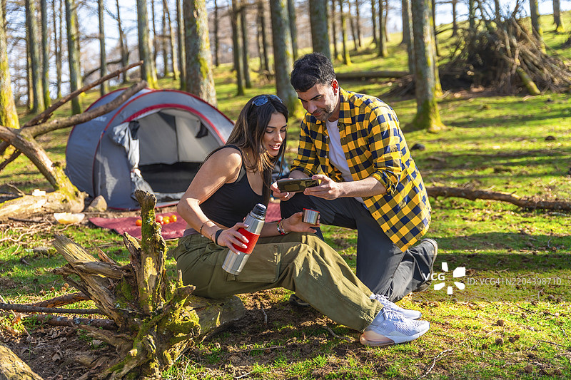 一对徒步旅行者在树林中享受露营之旅，边喝热饮边在智能手机上观看照片图片素材