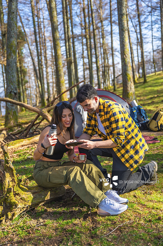 年轻情侣在阳光明媚的森林里露营，享用热饮，同时使用平板电脑，靠近他们的帐篷。图片素材