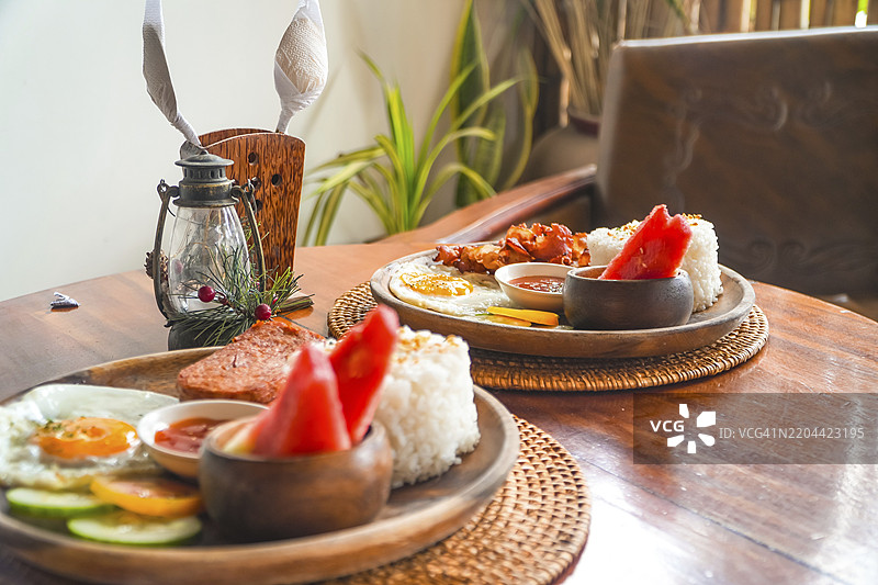 两盘菲律宾早餐，包含糖醋肉、炒饭、煎蛋、西瓜、黄瓜和番茄，摆放在菲律宾希亚高岛的木桌上。图片素材