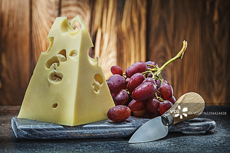 一块马斯达姆奶酪放在复古的切割板上，旁边有刀和一串玫瑰葡萄，背景是古老的木材。图片素材