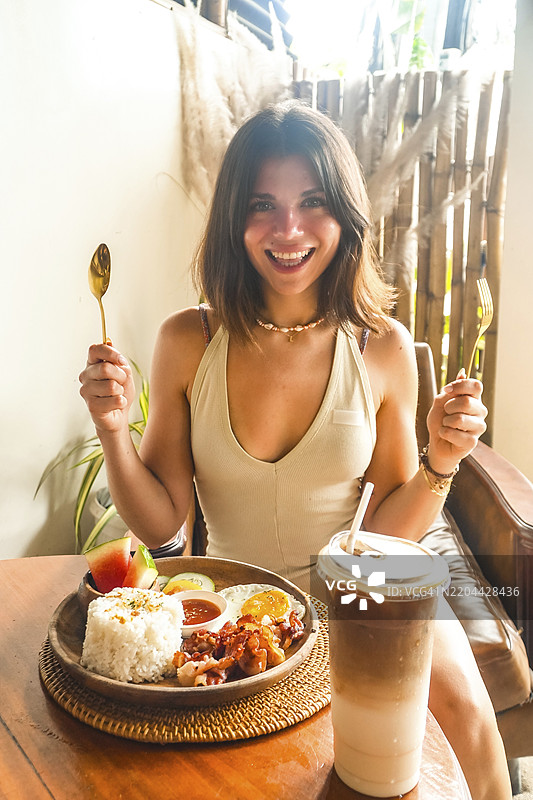 年轻女性手持餐具微笑，准备在菲律宾希亚高岛的一家餐厅享用传统菲律宾早餐和冰咖啡。图片素材